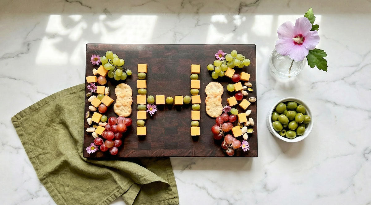 end grain cutting board with cubed cheese and green olives arranged in the letter 'H' with grapes and crackers surrounding on marble counter