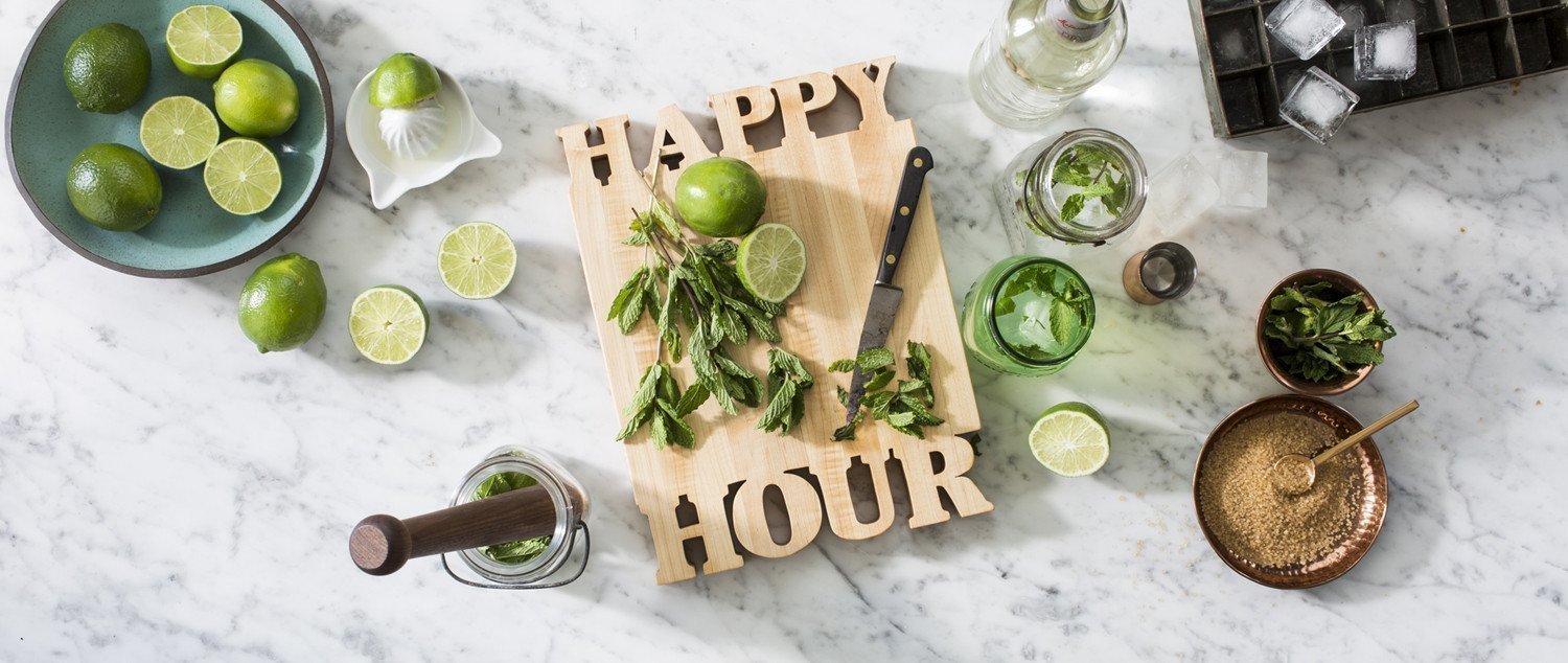 maple cutting board with 'Happy Hour' cut out of wood, mojito drink, limes, mint on marble background