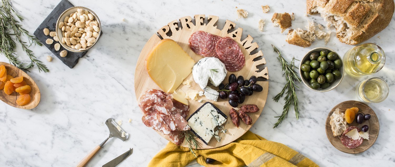 Round maple personalized cutting board with the name 'PEREZ FAMILY' cut out of the wood, charcuterie spread on marble background