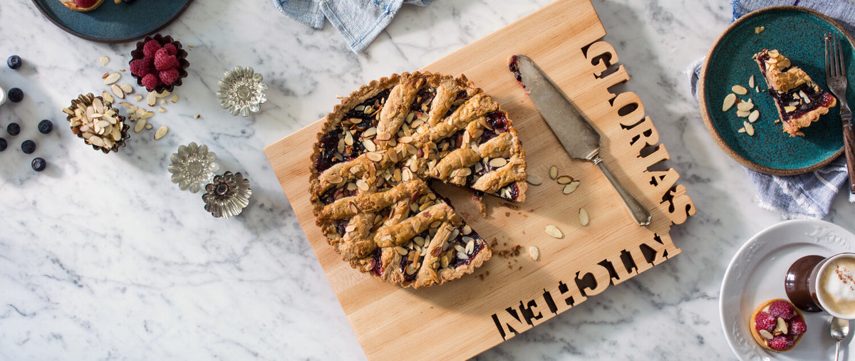 maple rectangle cutting board with 'Gloria's Kitchen' cut our of the top of the wood, shown with cherry pie on marble counter