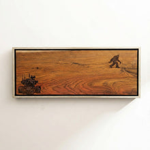 Wooden plaque with engraved images of a big foot and a vehicle framed on a white wall.