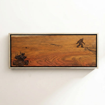 Wooden plaque with engraved images of a big foot and a vehicle framed on a white wall.