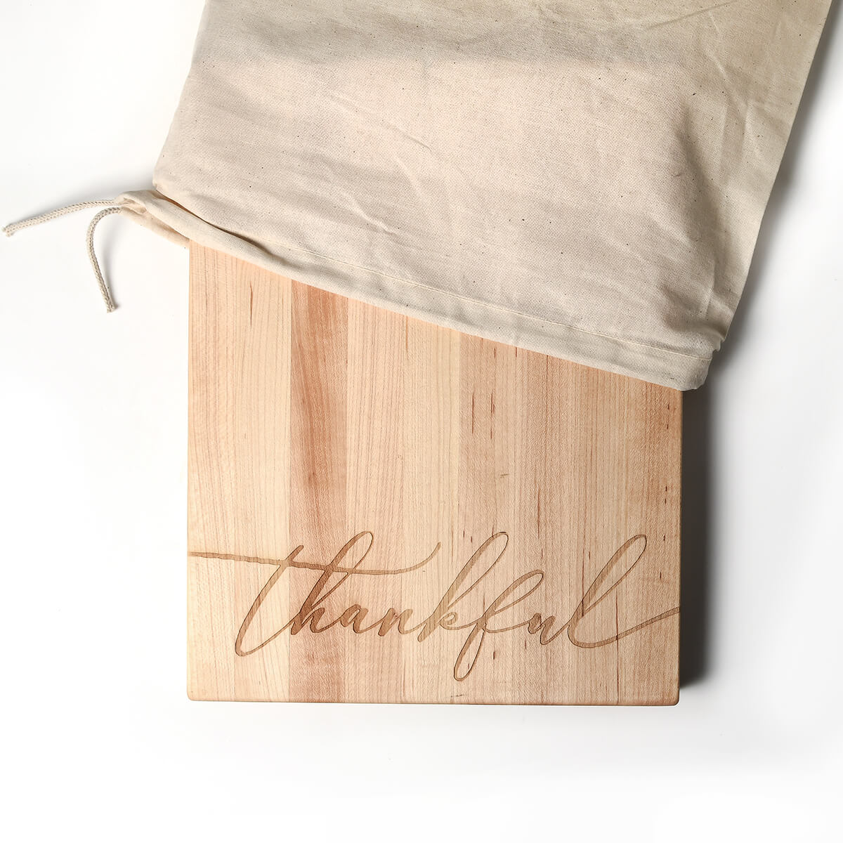 maple wood cutting board with the word 'thankful' laser engraved on the bottom with a cotton bag on white background