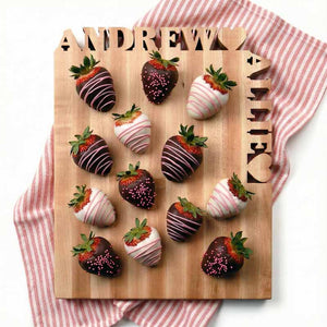 Wooden board with chocolate-dipped strawberries and first names cut out of the wood on a red and white striped cloth.