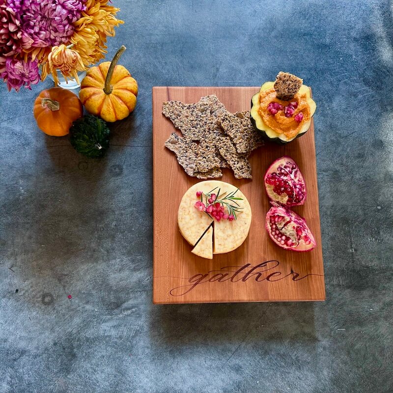 Wooden board with cheese, crackers, and pomegranate seeds on a textured surface with pumpkins and flowers.
