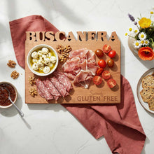 Wooden gluten free gifts charcuterie board with meats, cheese, and tomatoes on a light surface with 'Buscanera' cutout of top and 'Seriously Gluten free' laser engraved on lower right side..