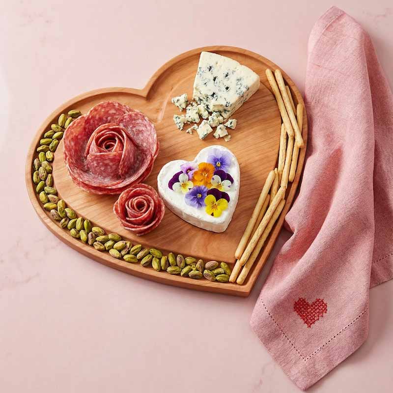 Heart-shaped cheese board with salami roses, cheese, and flowers on a pink background.