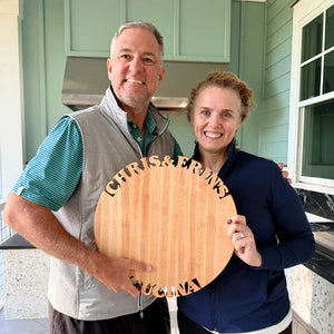 Two people holding a wooden lazy susan  with 'Chris & Erin's Cucina' cut out of the wood, standing in front of a house.