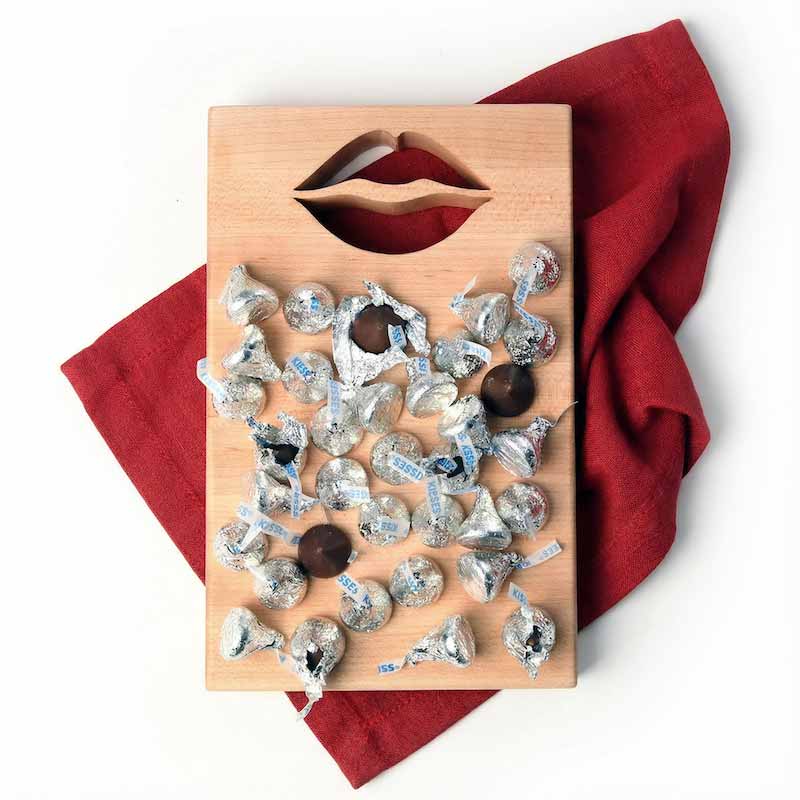 Wooden cutting board with lips shape cut out with chocolate kisses on top of board and the board is on a red cloth background