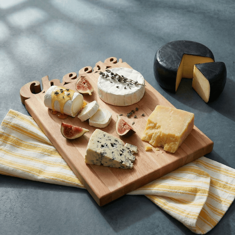 wooden cheese board showing 3 different cheeses and cut figs on the board and one cheese on the counter, board is on a gray counter with yellow and white towel