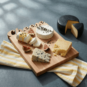 wooden cheese board showing 3 different cheeses and cut figs on the board and one cheese on the counter, board is on a gray counter with yellow and white towel