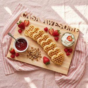 Wooden board with heart-shaped waffles, berries, and toppings on a pink cloth background. Board is personalized with the words 'casa awesome'.
