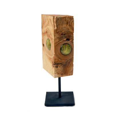 a piece of wood with 2 green reptile like eyes and its shown on a black metal pole and base
