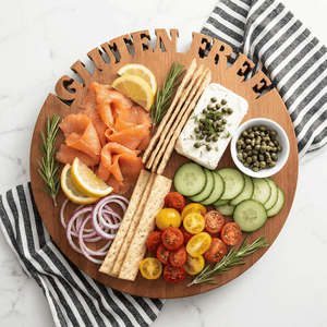 Round wooden cutting board with  'GLUTEN FREE' cut out of top, smoked salmon, vegetables, and crackers on a marble surface.
