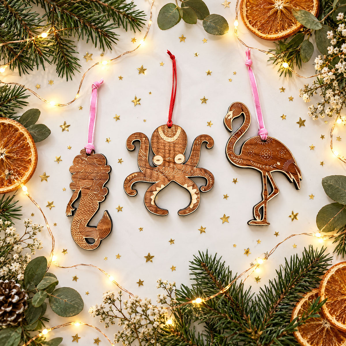 mermaid and octupus and flamingo shaped wooden holiday ornaments, on white backgrounded surrounded by gold glitter, greenery, fairy lights