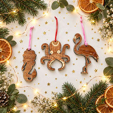 mermaid and octupus and flamingo shaped wooden holiday ornaments, on white backgrounded surrounded by gold glitter, greenery, fairy lights