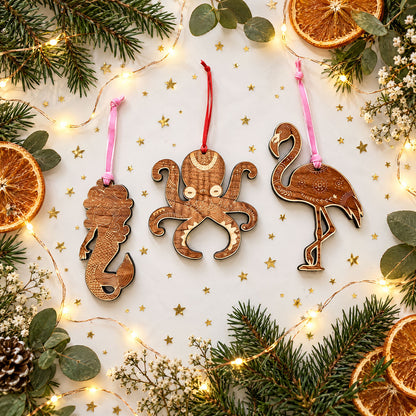 mermaid and octupus and flamingo shaped wooden holiday ornaments, on white backgrounded surrounded by gold glitter, greenery, fairy lights