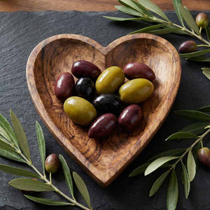 Heart-shaped wooden bowl filled with olives on a dark slate surface with olive branches.