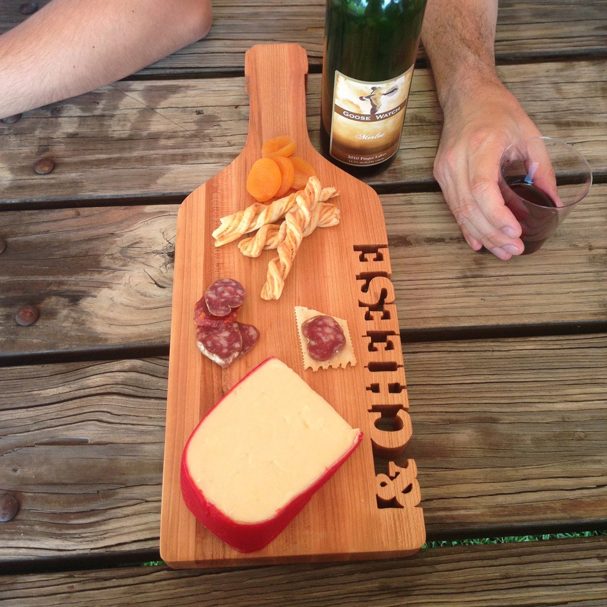 wine bottle shaped maple board with the words &cheese cut out, shown with cheese, crackers and a bottle of wine on picnic table